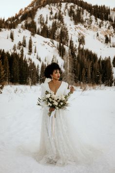 a woman standing in the snow wearing a wedding dress and holding a bridal bouquet