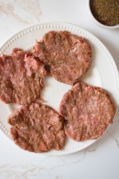 four hamburger patties on a white plate next to a bowl of seasoning