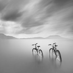 black and white photograph of two bicycles in the water with mountains in the distance behind them