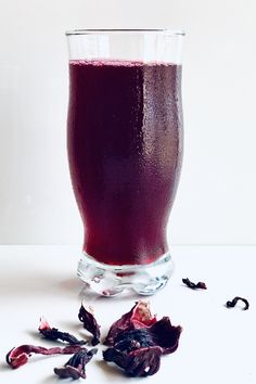 a glass filled with red liquid next to dried rose petals on a white counter top