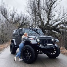 a woman sitting on top of a black jeep in the middle of a road next to trees