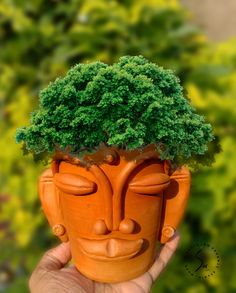 a hand holding up a clay face with a tree on it's head in front of some bushes