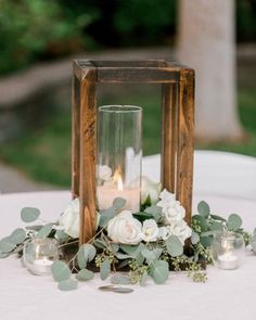 an arrangement of white flowers and greenery is displayed in a wooden lantern on top of a table