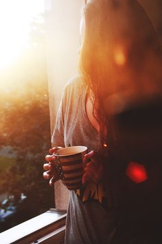 a woman holding a coffee cup in front of a window with the sun shining through