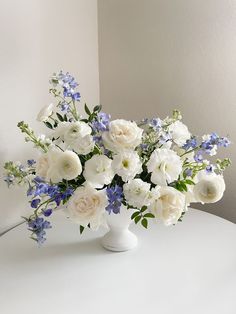 a white vase filled with lots of white and blue flowers on top of a table