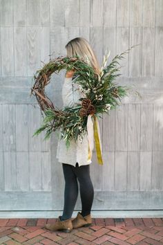 a woman holding a wreath with pine cones and greenery in front of a wall