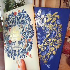 a hand holding an open book in front of a blue and yellow book with gold foil on it