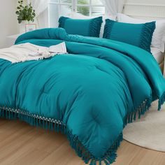 a teal colored comforter with tassels on the edges and pillows, in front of a window