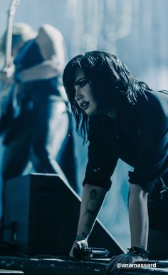 a woman with long hair and piercings on her arm, bending over in front of a stage