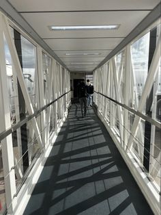 two people walking down a long white walkway with metal railings on both sides and sun shining through the windows