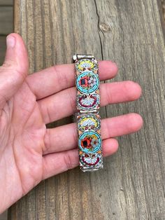 This bracelet has 5 wide rectangular segments covered in beautifully designed micro mosaic patterns. Each segment has a circle in the center filled with an orange and blue flower surrounded by a turquoise border. Some of the flower clusters also have a tiny yellow flower tucked in on the side. The pattern is outlined in twisted silver metal borders and the back of the bracelet is a matte silver tone. There is minor wear, specifically on the metal back but overall the bracelet is in very good con Bohemian Round Meenakari Bracelets, Artisan Multicolor Bracelets With Inlay, Multicolor Artisan Bracelet With Inlay, Bohemian Square Multicolor Jewelry, Multicolor Artisan Inlay Bracelets, Multicolor Wearable Art Bracelet, Adjustable Multicolor Inlay Bracelets, Multicolor Inlay Cuff Bracelet Gift, Rectangular Multicolor Bracelet