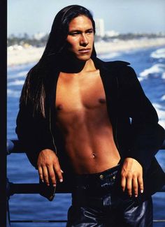 a man with long hair and no shirt standing next to the ocean wearing leather pants