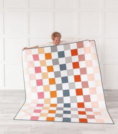 a man holding up a large quilt in front of a white wall with an orange and grey checkerboard pattern