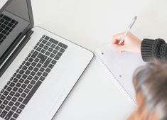 a person writing on a notebook next to a laptop