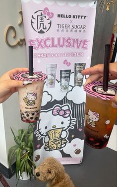 two people holding up hello kitty drinks and a teddy bear sitting on the table next to them