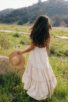 a girl in a white dress and straw hat walking through the grass with her back to the camera