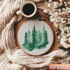 a cross - stitch pattern with green trees on it next to a cup of coffee