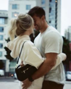 a man and woman walking down the street in front of tall buildings with their arms around each other