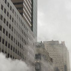 an airplane is flying in the air near tall buildings and steam rises from the ground