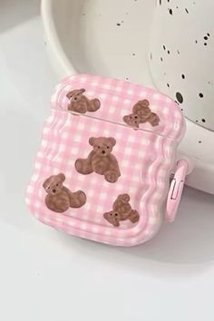 an apple airpods case with teddy bears on pink checkered fabric, sitting next to a white bowl