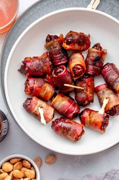 bacon wrapped dates on skewers in a white bowl next to nuts and drinks