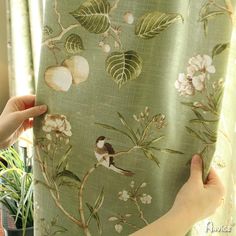 two hands are holding up a curtain with birds and flowers on it in front of a potted plant