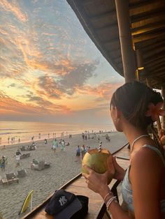 a woman sitting at a table on top of a beach next to the ocean holding a coconut