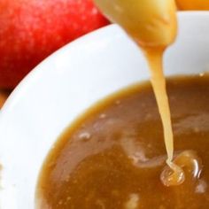 someone pouring caramel sauce into a white bowl with apples and oranges in the background