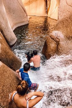 three people are sitting in the water near some rocks and one person is standing up