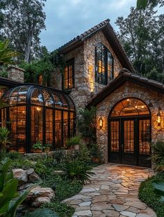 a stone and glass house surrounded by greenery at night with lights on the windows