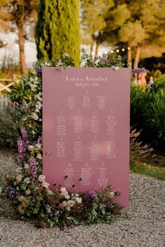 a pink sign with flowers on it sitting in front of some bushes and trees at an outdoor event