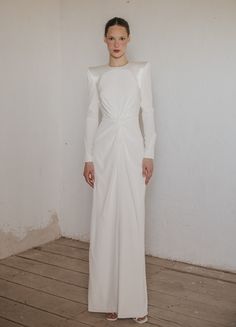 a woman standing in front of a white wall wearing a long white dress with an asymmetrical neckline