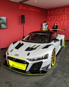 two people standing on the roof of a white sports car in a red room with an advertisement behind it