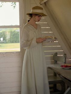 a woman in a white dress and straw hat is reading a book near a window