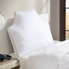 a bed with white sheets and pillows next to a lamp on a table in front of a window