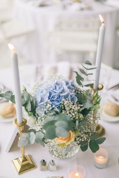 a centerpiece with blue flowers and greenery sits on a white tablecloth surrounded by candles