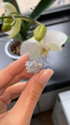 a woman's hand holding an engagement ring in front of a vase with flowers