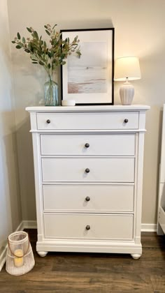 a white chest of drawers with flowers on top next to a lamp and framed photograph