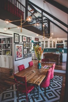 a dining room table with red chairs and pictures on the wall above it in front of a spiral staircase