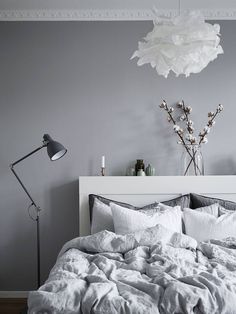 a bed with white sheets and pillows next to a lamp on a table in front of a gray wall