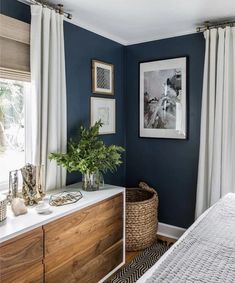 a bedroom with dark blue walls and white drapes on the windowsill, a large wooden dresser has a plant in front of it