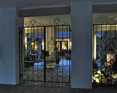 an iron gate in front of a house at night with the lights on and plants outside