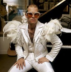 a man in white suit and sunglasses sitting next to a piano with his hands on his hips