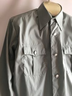 Awesome vintage men's uniform work shirt jacket. The military inspired cut and color encourage me to believe this was a aviation uniform of some sort. Somewhere between a shirt and a jacket, these piece has some great details. The material is most likely a permanent press style poly/cotton blend, though no content label is present. The shirt has a button front styling much like a classic men's dress shirt but with a more boxy cut and elastic waist. It has small loops and holes near top shoulder Military Style Collared Shirt For Workwear, Military Style Button-up Work Shirt, Military Style Button-up Shirt For Work, Military Style Shirt For Work With Button Closure, Military Long Sleeve Work Shirt, Military Style Workwear Button-up Shirt, Military Style Shirt With Buttons For Work, Military Style Collared Shirt With Snap Buttons, Military Style Workwear Shirt With Buttons