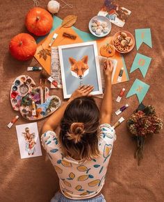 a woman sitting at a table with art supplies and pictures on the floor next to her