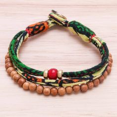 For an appealing addition to your wardrobe this bracelet from Thailand is created by Thanadol. He selects beads of petite sese wood strung along two strands of cotton printed with green and multicolored designs. One of the cotton strands is centered by a red glass bead that is flanked by two beads of brass. A brass button clasp closes this boho accessory. Adjustable Green Beaded Bracelets With Wooden Beads, Adjustable Green Bracelet With Wooden Beads, Bohemian Wooden Beaded Friendship Bracelets, Brass Pendant Necklace, Casual Bracelets, Fabric Bracelets, Wood Bracelet, Boho Accessories, Patchwork Fabric