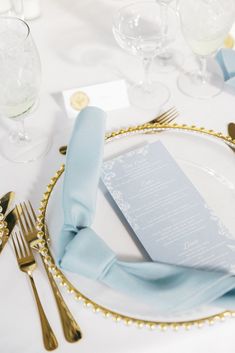 a place setting with blue napkins and gold rimmed cutlery on a white table cloth