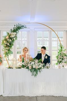 a bride and groom sitting at a table with greenery