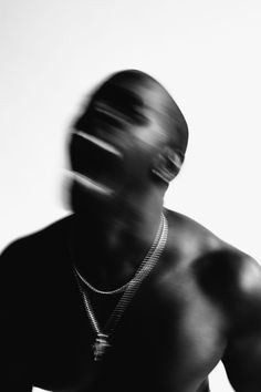 a man in black shirt and necklace standing next to a white wall with blurry image