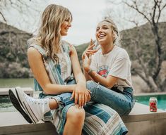 two young women sitting next to each other on a cement wall near a pool and trees